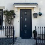 Elegant black XtremeDoor composite door with a minimalistic design, featuring rectangular glass panels, set within a white door frame, matched with black-framed windows, against a light grey exterior wall, secured by a black iron fence.