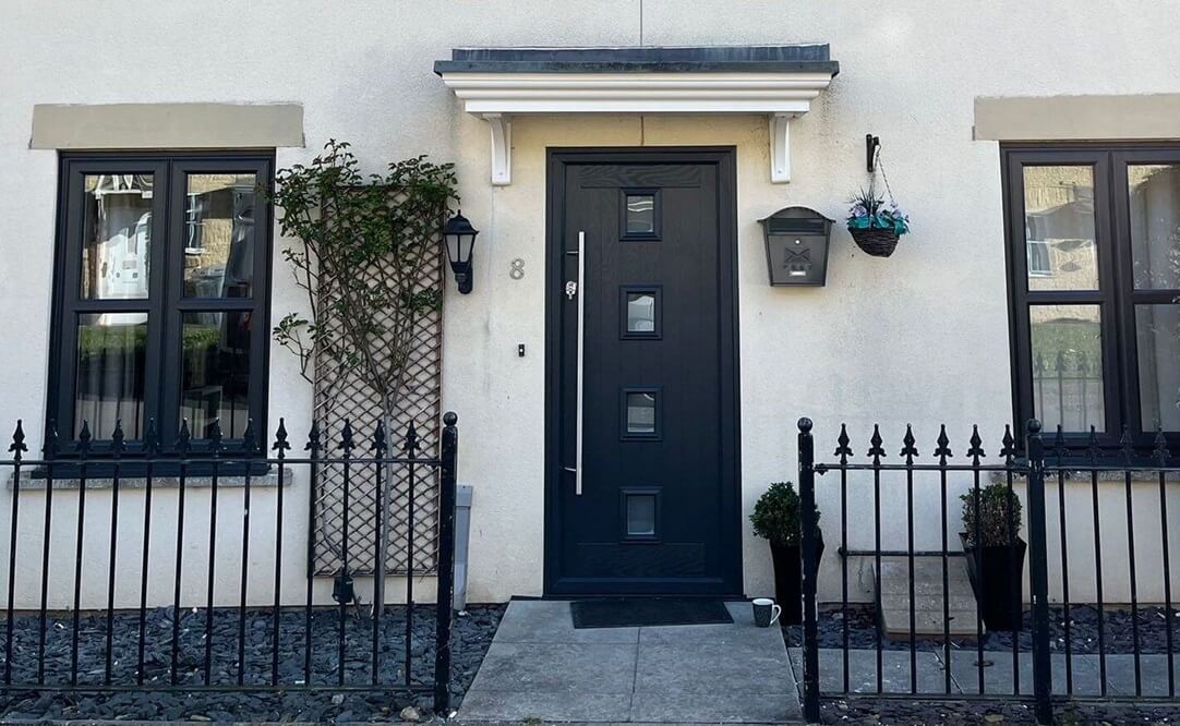 Elegant black XtremeDoor composite door with a minimalistic design, featuring rectangular glass panels, set within a white door frame, matched with black-framed windows, against a light grey exterior wall, secured by a black iron fence.